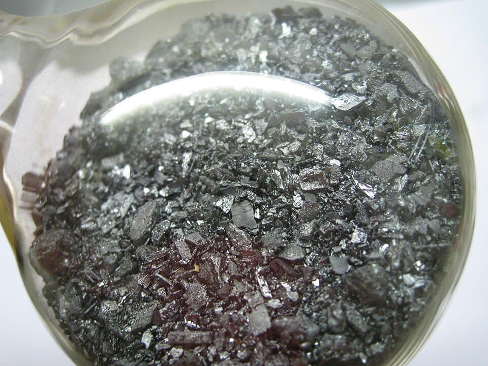 Dark grey metallic iodine crystals, 99.9% purity, in a glass flask.